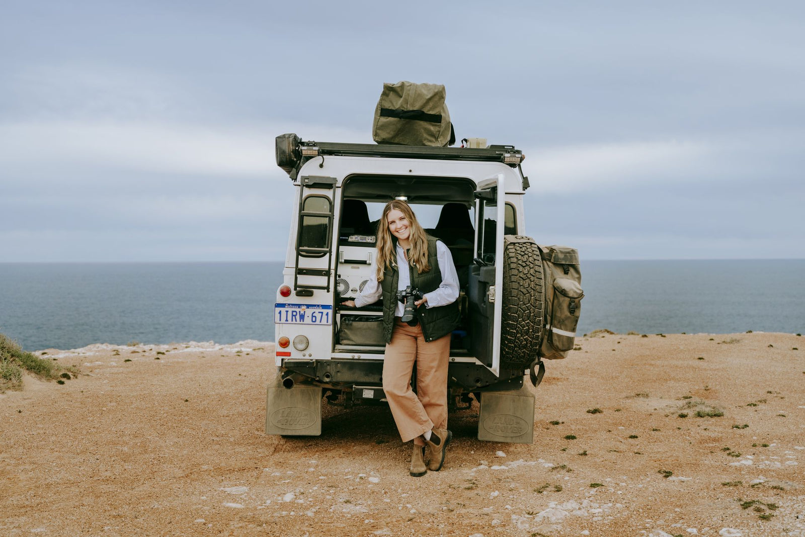 Woman leaning on the back of a Defender with a Down Under fridge freezer inside, on a clifftop above the ocean
